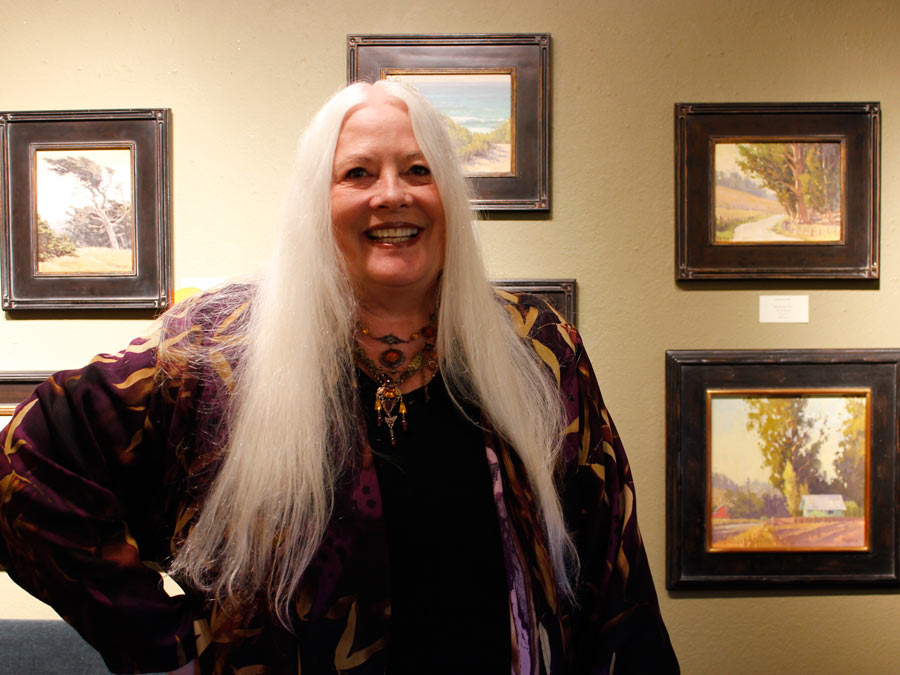 A woman with long white hair smiles in front of a yellow wall with paintings hanging on it