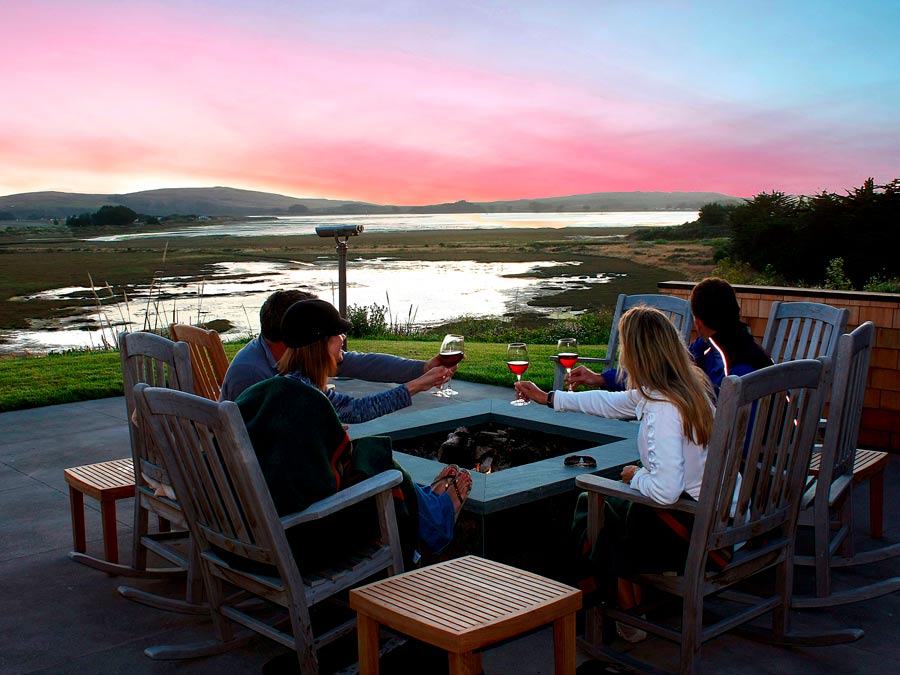 People toast with glasses of wine as the sun sets, sitting around a firepit