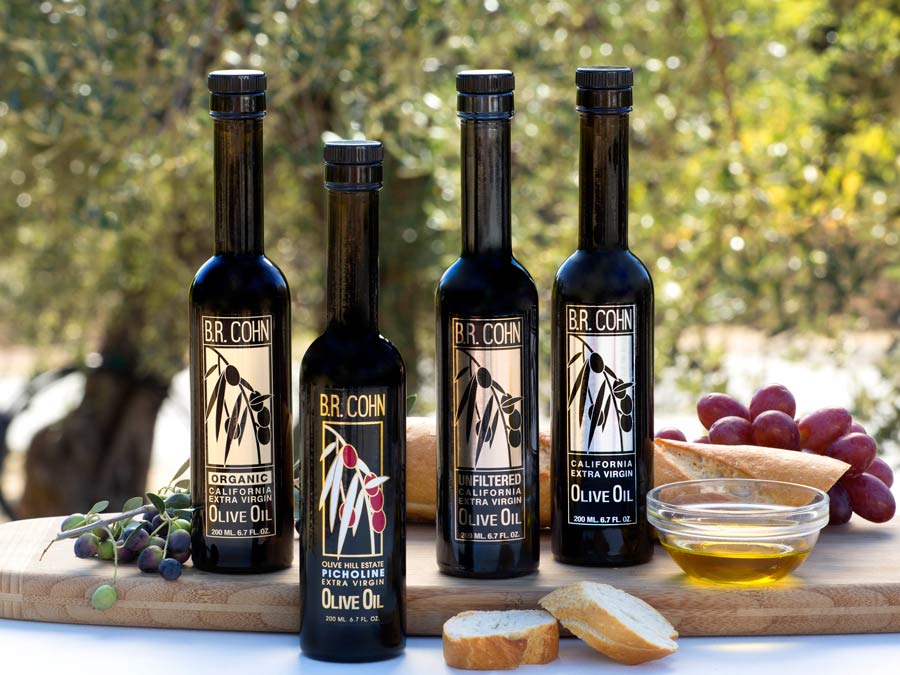 Bottles of olive lined up on a wooden platter with bread, olives, and purple grapes