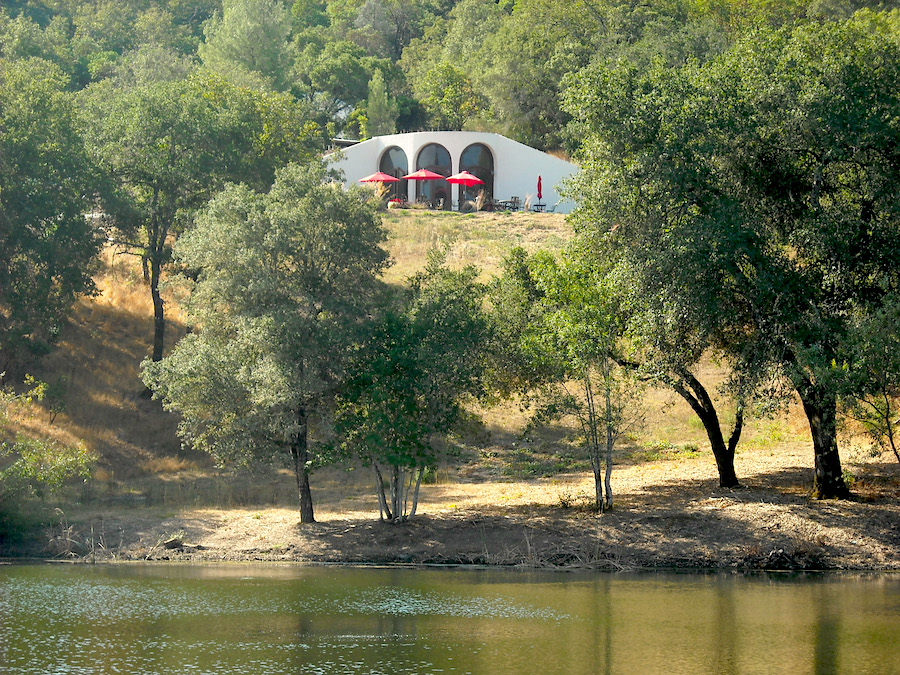 Curved, white winery building carved into a hillside, with arched windows in their tasting room, and red umbrellas on a sunny patio overlooking a green pond. 